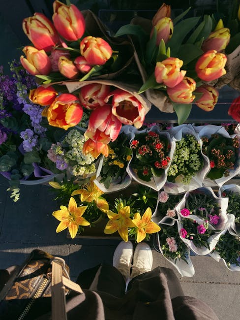 A top-down view of a variety of fresh flower bouquets arranged outdoors, featuring vibrant tulips with yellow and orange petals, surrounded by smaller floral arrangements in shades of purple, red, yel