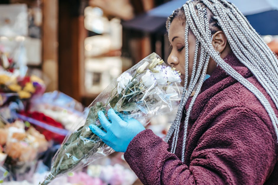A person with long, braided silver hair wearing a maroon fleece jacket and blue gloves is holding a clear plastic-wrapped bouquet of white roses and greenery. They are inspecting the fragrant, fresh f